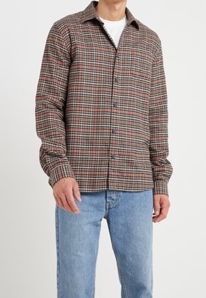 Uomo che indossa una camicia a quadri marrone, nera e arancione sopra una t-shirt bianca, abbinata a jeans azzurri, in piedi contro uno sfondo bianco.