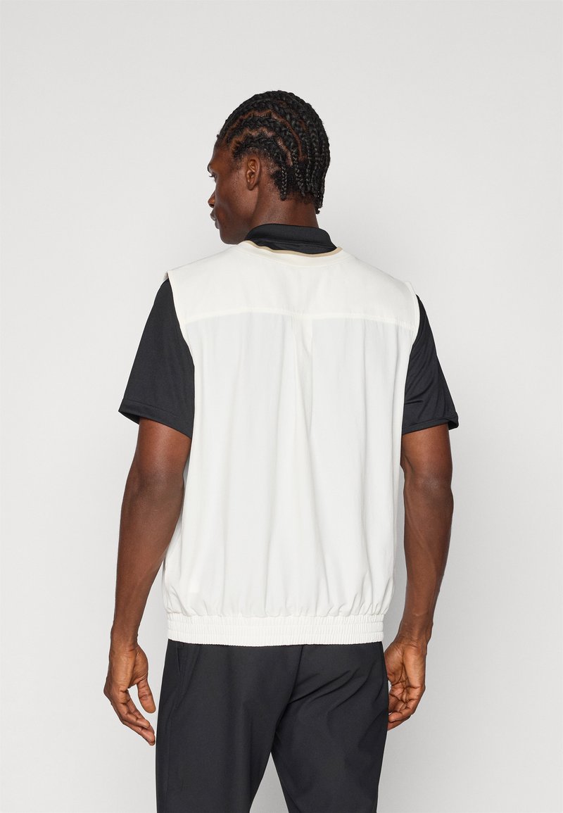 White sleeveless vest with elastic bottom, contrasting black collar, and back section featuring a smooth texture. Worn over a black shirt.