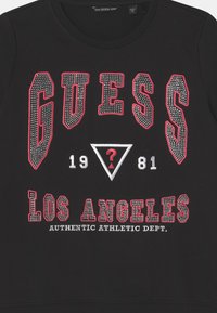 Felpa nera con la scritta "GUESS" e "LOS ANGELES" in rosso, dettagli in strass, una grafica triangolare e testo bianco sottostante.