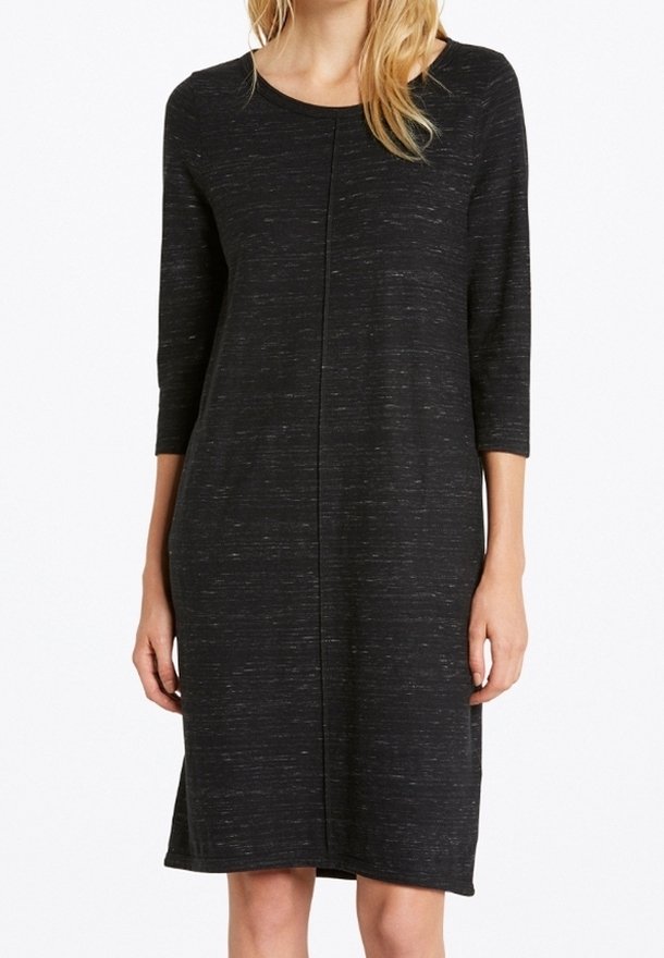 Femme portant une robe noire, longue aux genoux et à manches longues, avec une texture subtile et une couture centrale verticale à l'avant.