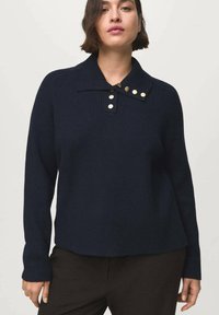 Donna che indossa un maglione a coste blu scuro con un ampio colletto caratterizzato da cinque bottoni dorati, abbinato a pantaloni scuri.