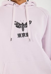 Lys rosa hettegenser med frontlomme, prydet med en svart møllgrafikk og japansk tekst. Mykt stoff, hette med snor og metallspisser.