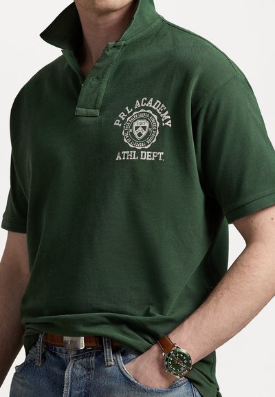 Cămașă polo verde cu guler clasic, având text imprimat alb și un logo cu scut pe piept. Material din bumbac cu finisaj texturat.
