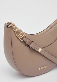 Sac en cuir taupe doux, forme arrondie avec une fermeture éclair et des accessoires dorés. Bandoulière réglable et logo embossé sur le devant.