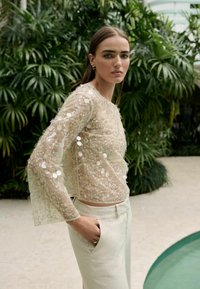 Haut beige translucide avec un délicat brodé floral blanc et des embellissements circulaires. Associé à un pantalon en lin de couleur claire, sur fond de verdure.