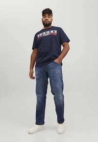 Navy zila T-krekls ar uzrakstu "JACK & JONES", kopā ar ziliem džinsa džinsiem un baltām sporta kurpēm. Modelis stāv ar roku kabatā pret vienkāršu fonu.