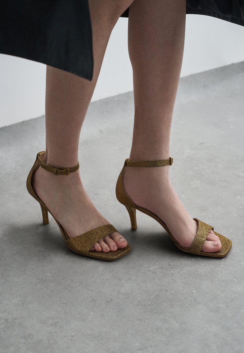 Sandalias de tacón con glitter dorado, con puntera cuadrada, tiras abiertas y una correa ajustable en el tobillo. Presenta un fino tacón de aguja.