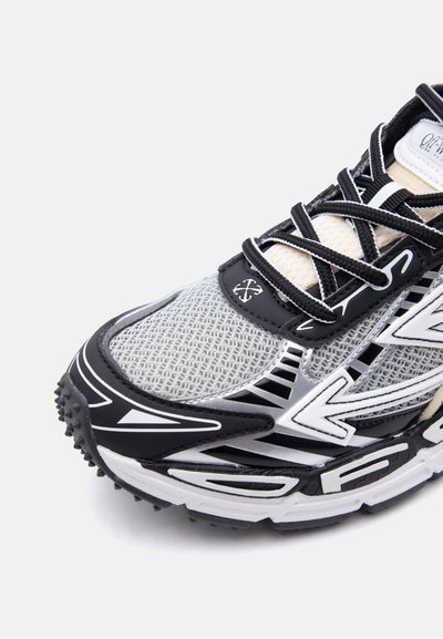 Αθλητικό παπούτσι με μαύρο, λευκό και ασημί σχεδιασμό από δίχτυ, μαύρα κορδόνια, υφή στη σόλα και λογότυπο σε σχήμα βέλους στο πλάι.