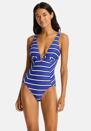 Femme portant un maillot de bain une pièce rayé bleu, blanc et noir avec un profond décolleté en V et un fermoir doré au centre devant.