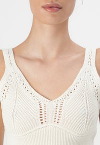 Elfenbenfarget strikket tank topp med V-hals, tykke stropper og teksturerte mønstre med åpne masker. Myk stoff med tettsittende design.