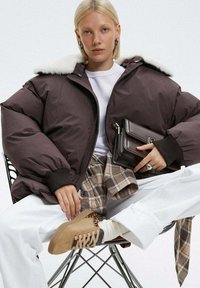 Brązowa kurtka puffar z białym sztucznym futrem w kołnierzu, zestawiona z białą bluzą, w kratkę spodniami i beżowymi sneakersami z akcentami w motyw zwierzęcy.