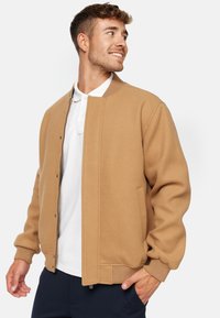 Khaki bomber bunda se zipovým uzávěrem, žebrovaným límcem a manžetami, bočními kapsami, vyrobená z měkkého texturovaného materiálu. Nositá přes bílou polo košili.