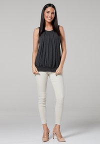Dark gray sleeveless top with pleated design and rounded hem, paired with cream skinny jeans. Model wearing nude high-heeled shoes.