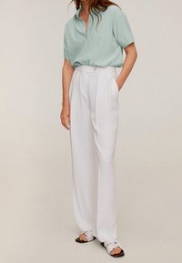 Femme portant une chemise à manches courtes vert clair, boutonnée, rentrée dans un pantalon large blanc taille haute, avec des sandales.