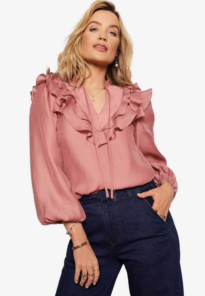 Rózsaszín blúz, amelynek nyakrészén réteges fodrok találhatók, hosszú puffos ujjak és megkötő rész, sötétkék farmernadrággal párosítva.