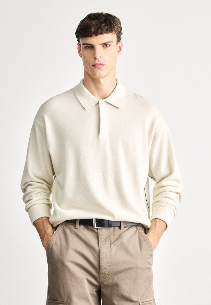 Κρεμ πολο πουλόβερ με πλεκτό σχέδιο και γιακά, χαλαρή εφαρμογή, ριγέ μανσέτες και διακριτή υφή, σε συνδυασμό με χακί cargo παντελόνια και μαύρη ζώνη.