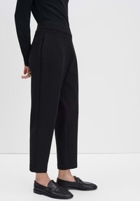 Zwarte tailored broeken met een hoge taille, zijzakken en een recht vallende pasvorm. Glad textuur en strakke lijnen. Gecombineerd met zwarte loafers.