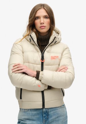Beige donsjacket met een getailleerde silhoutte, capuchon, zwarte rits en oranje logo. Gebreide donkerbruine kraag en manchetten zichtbaar.