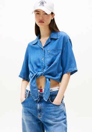 Camicia in denim azzurro chiaro con maniche corte, tasca frontale e vita annodata. Abbinata a jeans in denim a vita alta con una cintura che presenta il logo del marchio.