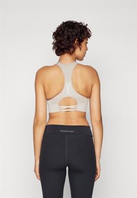 Jasno beżowy stanik sportowy z plecami w stylu racerback, z wycięciem i krzyżującymi się detalami z tyłu. Połączony z czarnymi legginsami o wysokim stanie.