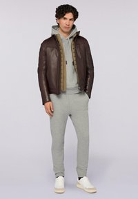 Kožená bomber bunda v tmavě hnědé barvě přes šedou mikinu, doplněná o světle šedé tepláky a bílé tenisky, s hladkou texturou a čistými liniemi.