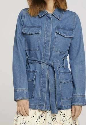 Kobieta nosząca niebieską kurtkę jeansową z czterema kieszeniami z patkami i związanym paskiem na białej spódnicy z czarnymi i brązowymi plamkami.