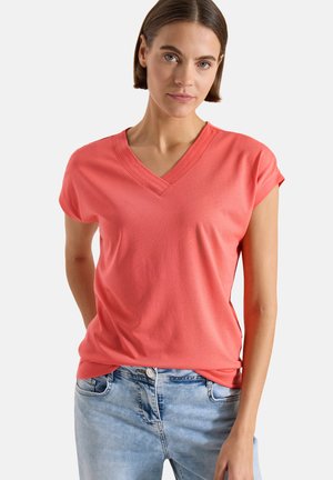 Kvinde iført koralrosa V-hals kortærmet top og lyseblå jeans, stående mod en ensfarvet lys grå baggrund.