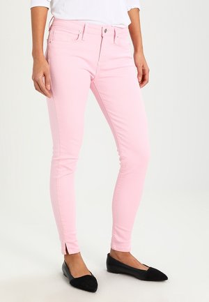 Osoba nosząca jasnoróżowe wąskie dżinsy z przednimi kieszeniami i rozcięciami przy kostkach, zestawione z czarnymi, szpiczastymi balerinami i białą bluzką.