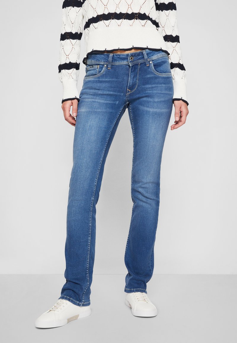 Jeans en denim bleu avec une coupe slim, dotés de poches avant, d'un léger délavage et de détails de couture, assortis à des baskets blanches.