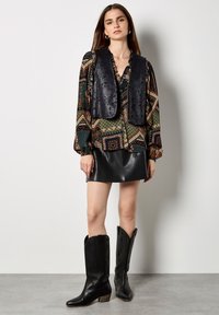 Gilet in pelle sintetica nera sopra una blusa a fantasia con tonalità di verde, arancione e marrone, abbinato a una mini gonna in pelle nera e stivali alti fino al ginocchio.