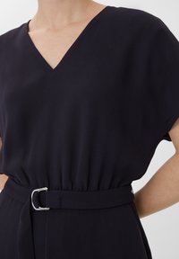 Kleid aus dunkelblauem Stoff mit V-Ausschnitt, kurzen Ärmeln, elastischem Bund und einem Gürtel mit einer silbernen Schnalle, der eine glatte Textur aufweist.