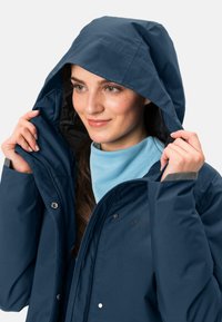 Giacca da pioggia blu navy con cappuccio, caratterizzata da una superficie liscia, bottoni a pressione e polsini regolabili, indossata sopra una maglietta azzurra chiara.