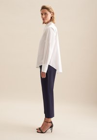 Weißes Langarmblusen mit spitzem Kragen, asymmetrischem Saum, kombiniert mit marineblauen schmal geschnittenen Hosen und schwarzen High-Heels.