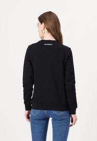 Svart sweatshirt med rund halsringning, ribbad mudd och nederkant. Har vitt logotyptext på ryggen. Bärs med blå jeans.
