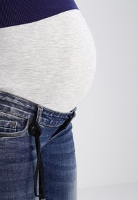 Äitiyspaita harmaan ja purppuran värimaailmalla, jossa on vatsabändi ja rento istuvuus, yhdistettynä sinisiin farkkuihin ja nyöreihin.