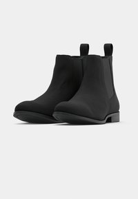 Paire de bottines noires à bouts ronds, panneaux élastiques latéraux, languettes à l'arrière et talons bas empilés sur un fond clair.