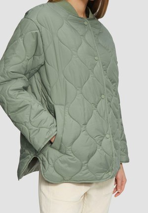 Personne portant une veste matelassée vert clair avec des boutons-pression et des poches, associée à un pantalon beige clair sur un fond uni.