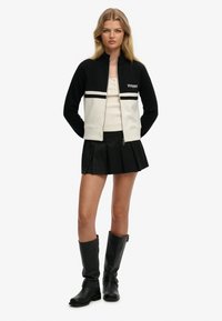 Czarna i biała kurtka na suwak z wysokim kołnierzem, ozdobiona poziomymi paskami i napisem „Sufferer”. Do tego plisowana czarna spódnica i kozaki za kolano.