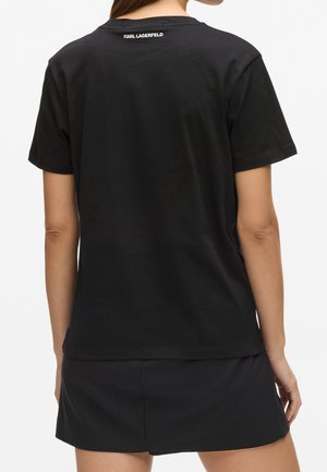 Nainen, jolla on yläselässä "KARL LAGERFELD" -teksti musta t-paita ja siihen sopiva musta hame, seisoo selin katsojaan.