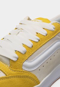 Κίτρινο και μπεζ αθλητικό παπούτσι με λευκά κορδόνια και λευκή πλευρική ρίγα, που δείχνει λεπτομερή υφή από σουέντ και διχτυωτό υλικό.