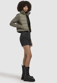 Vest de puf ușor, de culoare verde măsliniu, cu un guler înalt, combinat cu pantaloni scurți negri din denim și bocanci masivi negri cu șiret.