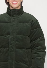 Chaqueta de plumas verde oscuro hecha de suave pana, con un alto cuello, secciones acolchadas y bolsillos laterales para uso práctico.