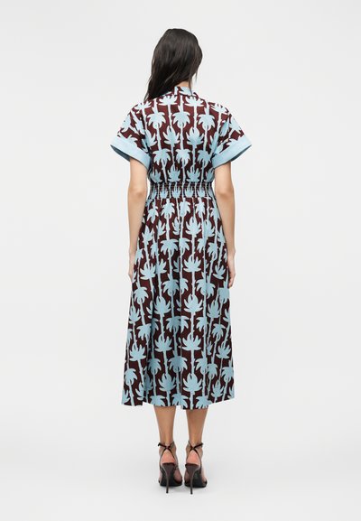 Γυναίκα στέκεται με την πλάτη, φοράει ένα καφέ μίντι φόρεμα με ανοιχτά γαλάζια σχέδια από φύλλα παλάμης, κοντά μανίκια, εφαρμοστή μέση και μαύρα σανδάλια με τακούνι.