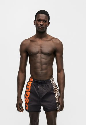 Athletic black man wearing black swim shorts with orange "DSQUA" lettering and leopard print panel standing against a plain light background.