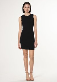 Schwarzes ärmelloses Kleid aus strukturiertem Stoff, figurbetontes Design und vertikale Nahtakzente an den Seiten. Getragen mit hellen High Heels.