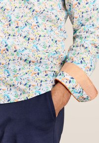 Bloemige shirt met een lichte achtergrond en kleurrijke bladerenpatronen, oranje accenten op de manchetten en een getailleerd ontwerp. Gekoeld met donkere broek.