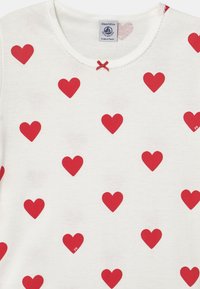 Top in cotone bianco con un motivo di cuori rossi. Collo tondo con bordino scallop e un piccolo fiocco rosso decorativo.