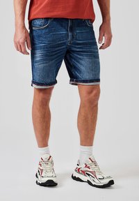 Shorts de denim en azul oscuro con desgastado, puños doblados y un forro rojo. Combinados con zapatillas blancas y negras y calcetines blancos.