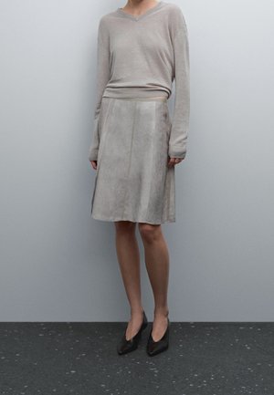 Personne portant un haut beige à manches longues avec col en V, une jupe gris clair longueur genou et des talons noirs à bout pointu, debout contre un mur gris.