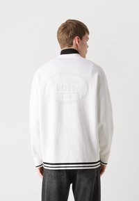 Suéter blanco con un collar negro y acentos de rayas. Presenta un sutil logo en relieve "BOSS" en la parte trasera. Textura de punto, corte relajado.